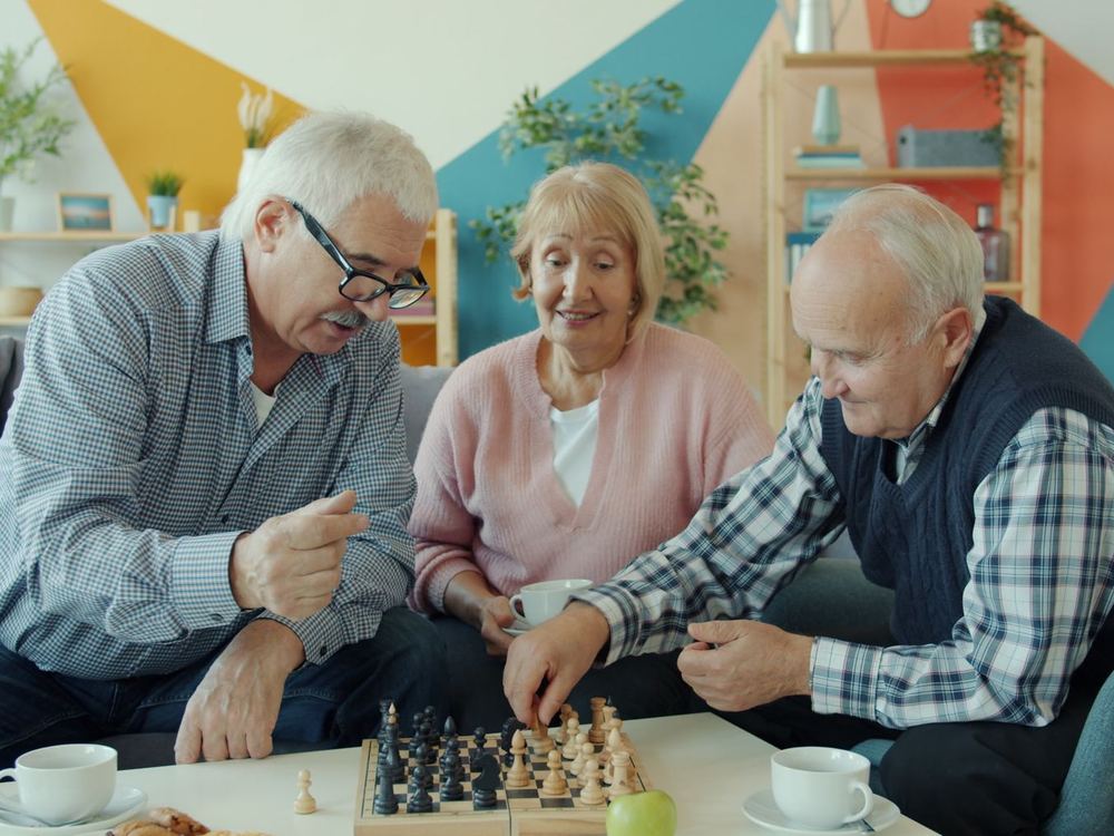 Three older adults laughing over a game of chess in a colorful living room