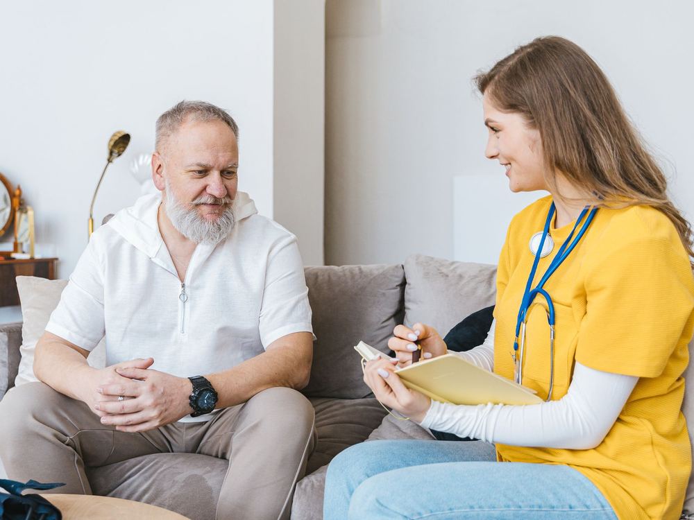 A nurse in scrubs talking with an older man on a couch at home
